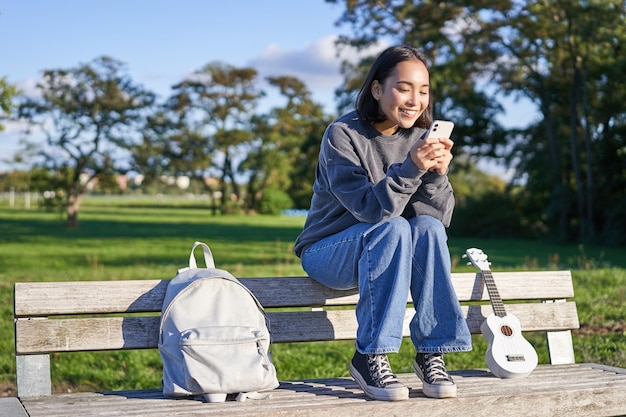 Mujer joven sentada en el parque en un banco con ukelele mirando el mensaje de lectura del teléfono inteligente en el ph móvil