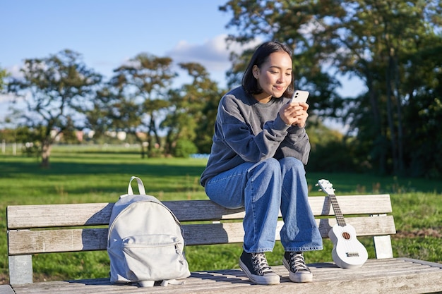 Mujer joven sentada en el parque en un banco con ukelele mirando el mensaje de lectura del teléfono inteligente en el ph móvil