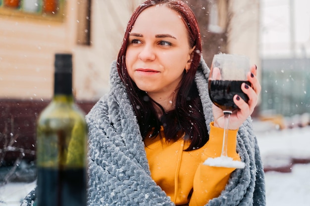 Mujer joven sentada en el patio y bebiendo vino tinto en tiempo de nieve Mujer envuelta en cuadros grises sentada en la calle con alcohol en temporada de invierno