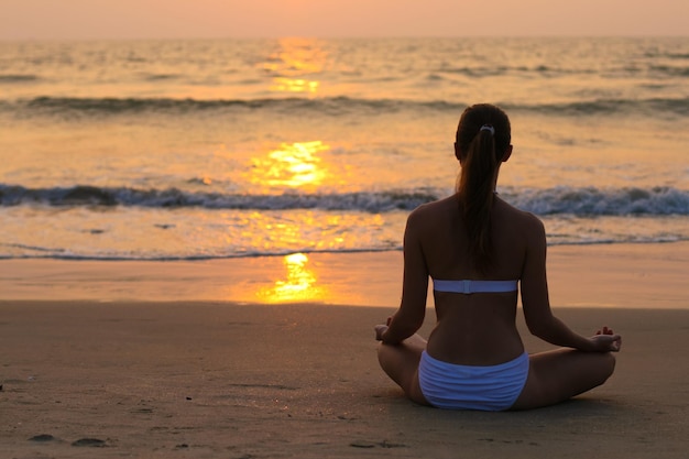 Mujer joven sentada en posición de loto en la playa al atardecer