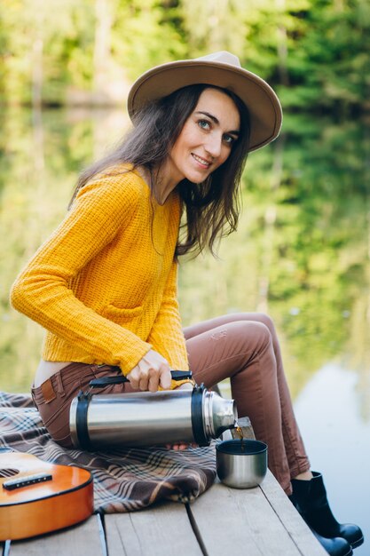 Mujer joven sentada en un puente sobre un lago con un paisaje otoñal y bebe té caliente de un termo. Viraje.