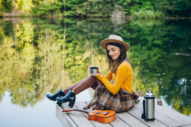 Mujer joven sentada en un puente sobre un lago con un paisaje otoñal y bebe té caliente de un termo. Viraje.