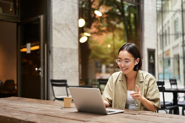 Mujer joven sentada en una reunión en línea en un café al aire libre hablando con una cámara portátil explicando algo