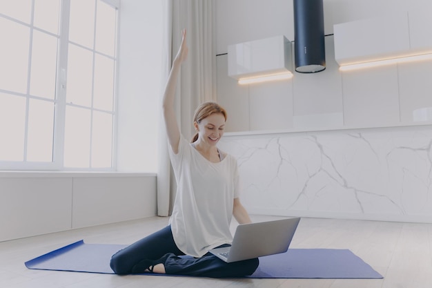 Mujer joven sentada de rodillas en el piso con una computadora portátil Tutorial de fitness en línea Entrenamiento a distancia