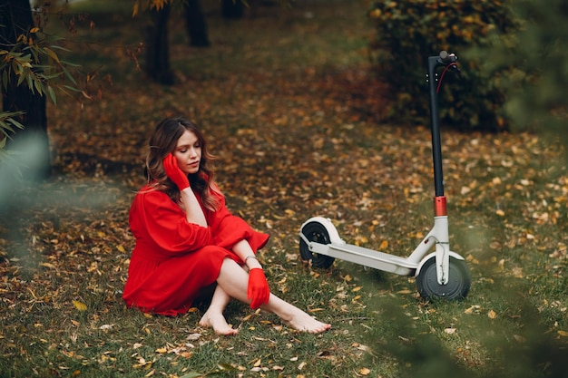 Mujer joven sentada con scooter eléctrico en vestido rojo en el parque de la ciudad de otoño