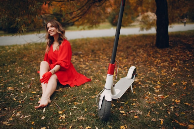 Mujer joven sentada con scooter eléctrico en vestido rojo en el parque de la ciudad de otoño