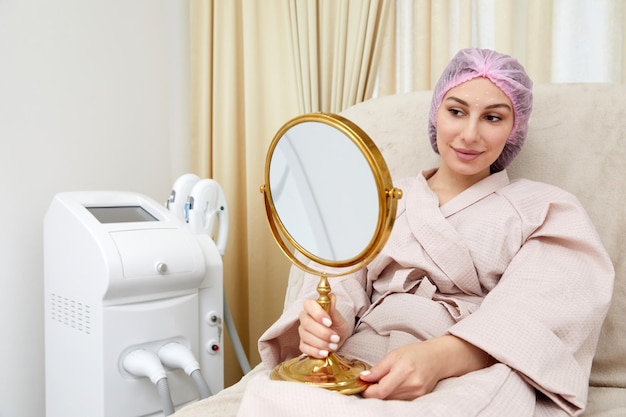 Mujer joven sentada en una silla médica y mirándose en el espejo Está satisfecha después de un tratamiento de belleza exitoso