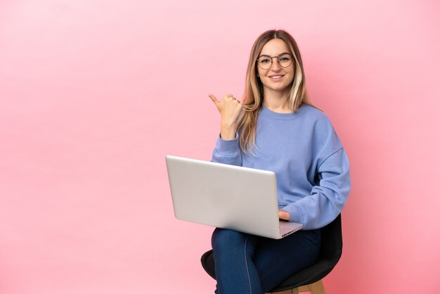 Mujer joven sentada en una silla con portátil sobre fondo rosa aislado con gesto de pulgar hacia arriba y sonriente