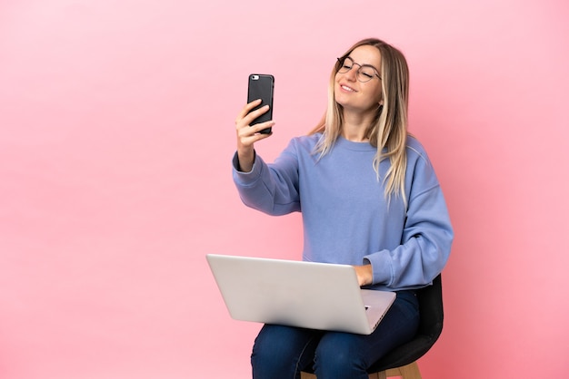 Mujer joven sentada en una silla con un portátil sobre fondo rosa aislado haciendo un selfie