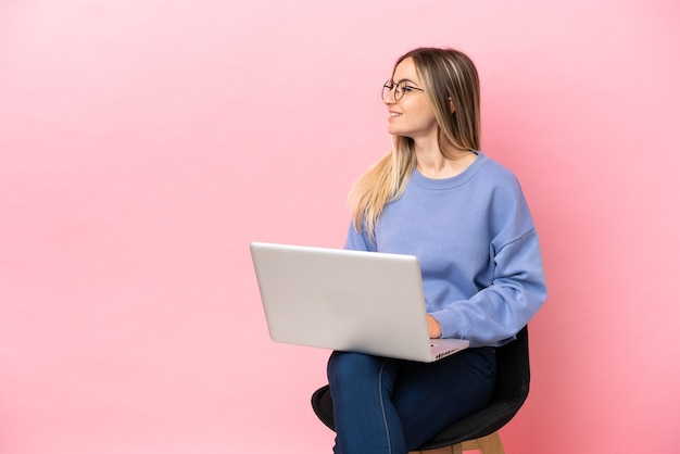 Mujer joven sentada en una silla con un portátil sobre fondo rosa aislado mirando hacia el lado