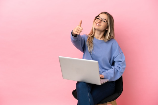 Mujer joven sentada en una silla con un portátil sobre una pared rosa aislada con los pulgares hacia arriba porque ha sucedido algo bueno