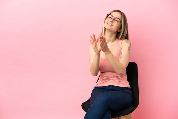 Mujer joven sentada en una silla sobre fondo rosa aislado aplaudiendo después de la presentación en una conferencia