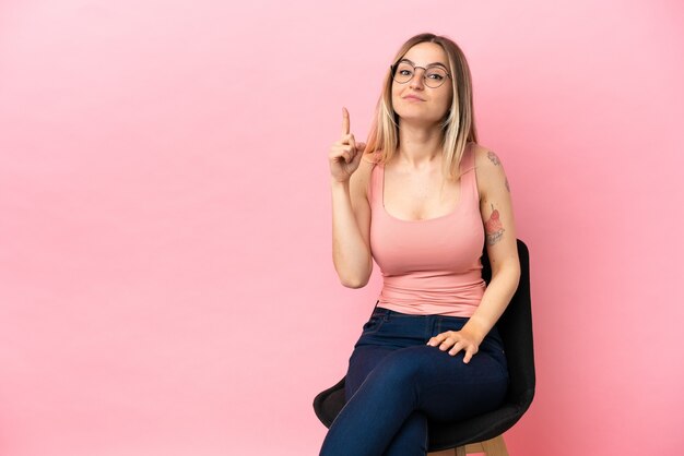 Mujer joven sentada en una silla sobre fondo rosa aislado apuntando con el dedo índice una gran idea