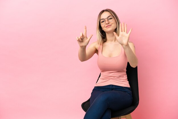 Mujer joven sentada en una silla sobre fondo rosa aislado contando siete con los dedos