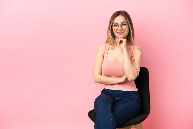 Mujer joven sentada en una silla sobre fondo rosa aislado con gafas y sonriendo