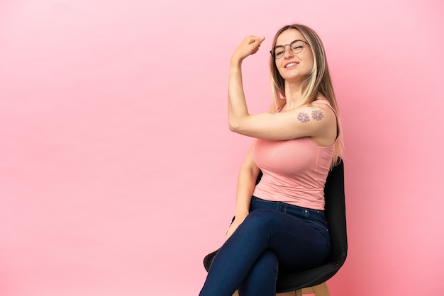 Mujer joven sentada en una silla sobre fondo rosa aislado haciendo gesto fuerte