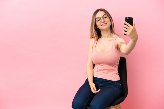 Mujer joven sentada en una silla sobre fondo rosa aislado haciendo un selfie