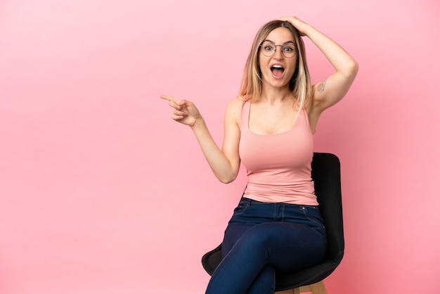 Mujer joven sentada en una silla sobre fondo rosa aislado sorprendido y apuntando con el dedo hacia el lado