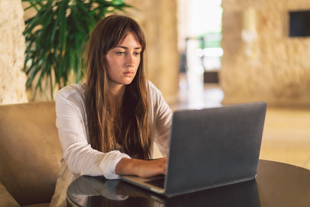 Mujer joven sentada en un sillón en el pasillo y trabajando en la computadora portátil