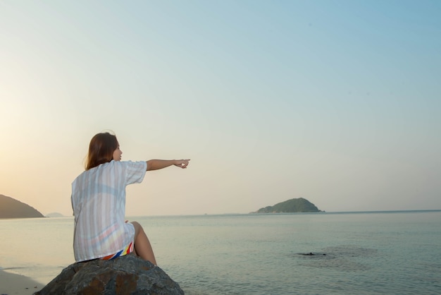 Mujer joven sentada sobre una roca disfrutando de la playa y apuntando al cielo