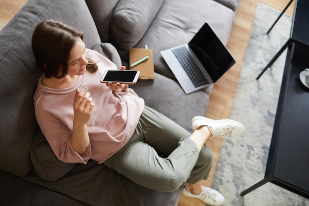 Mujer joven sentada en el sofá con una computadora portátil y hablando por teléfono móvil, ella trabaja en casa