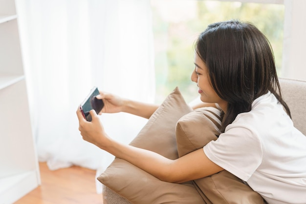 Mujer joven sentada en el sofá y jugando juegos en el teléfono inteligente en casa