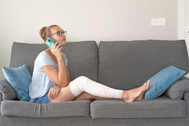 Mujer joven sentada en un sofá con una pierna rota en un vendaje hablando por teléfono móvil con la compañía de seguros