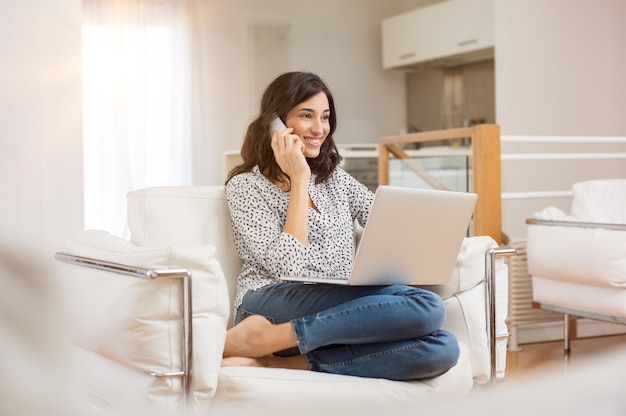 Mujer joven sentada en el sofá con el portátil y hablando por teléfono en casa