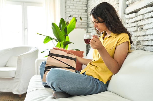 Una mujer joven sentada en el sofá saboreando una taza de té y eng