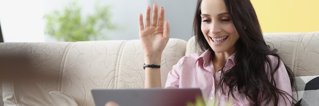 Mujer joven sentada en el sofá y saludando a la pantalla del portátil. Comunicación remota en concepto de redes sociales