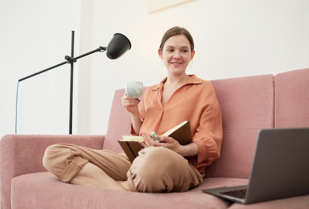 Mujer joven sentada en un sofá con una taza de café estudiando en línea en una laptop y tomando notas en un cuaderno en la habitación