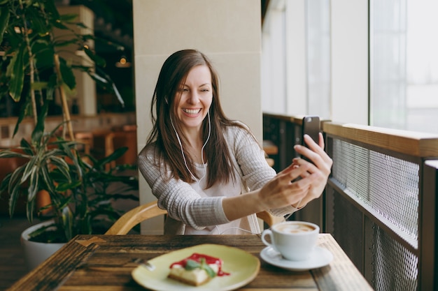 Mujer joven sentada sola en la cafetería a la mesa con una taza de capuchino, pastel, relajándose en el restaurante durante el tiempo libre. Mujer joven haciendo selfie en teléfono móvil, descansar en la cafetería. Concepto de estilo de vida.