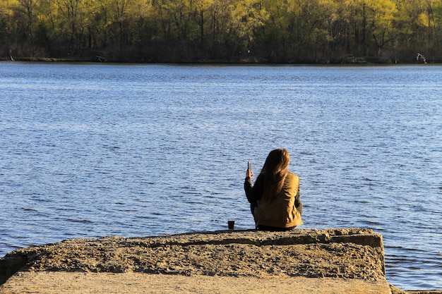 Mujer joven sentada sola en el muelle con taza de café. Vista trasera