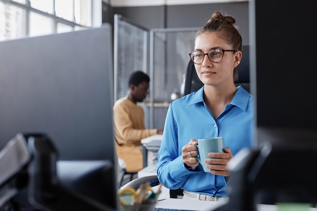 Mujer joven sentada en su lugar de trabajo y mirando el monitor de la computadora mientras bebe café