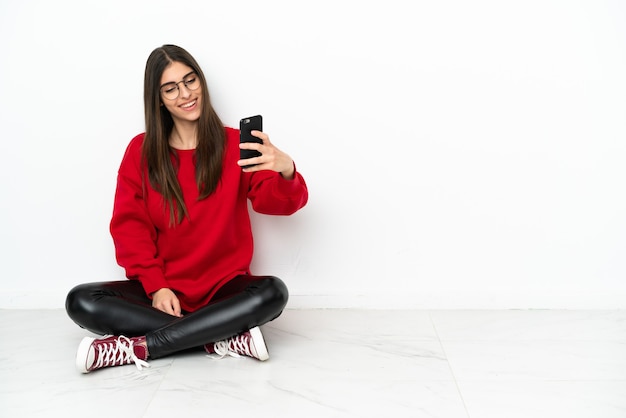 Mujer joven sentada en el suelo aislado sobre fondo blanco haciendo un selfie