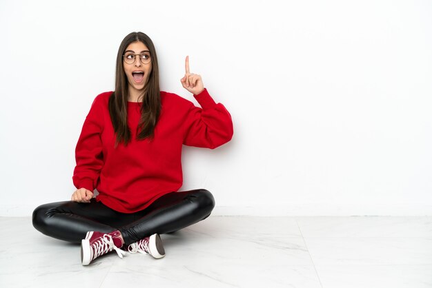 Mujer joven sentada en el suelo aislado sobre fondo blanco con la intención de darse cuenta de la solución mientras levanta un dedo