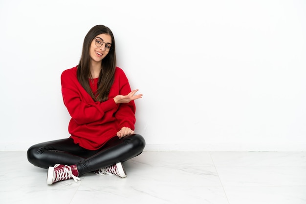 Mujer joven sentada en el suelo aislado sobre fondo blanco presentando una idea mientras mira sonriendo hacia