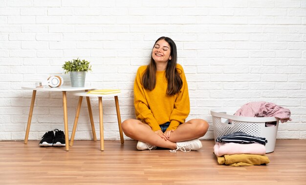 Foto mujer joven sentada en el suelo en el interior con cesto de ropa riendo