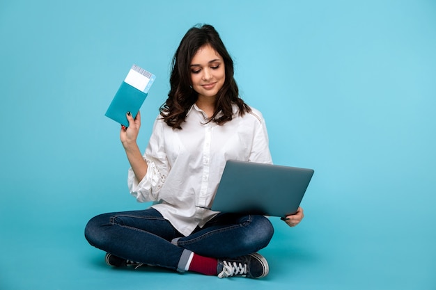Mujer joven sentada en el suelo con ordenador portátil y pasaporte comprando billetes online.