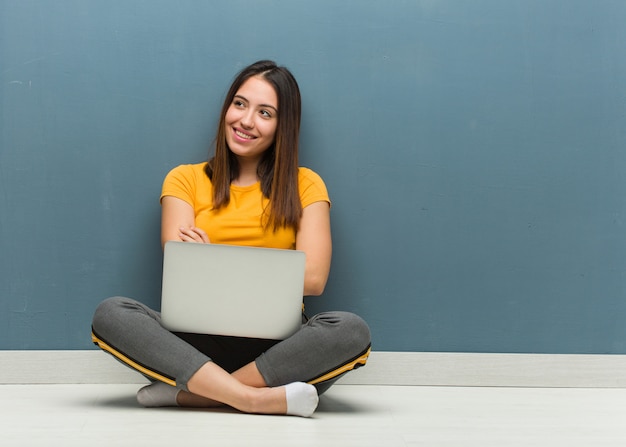 Mujer joven sentada en el suelo con un portátil sonriendo confiados y cruzando los brazos, mirando hacia arriba