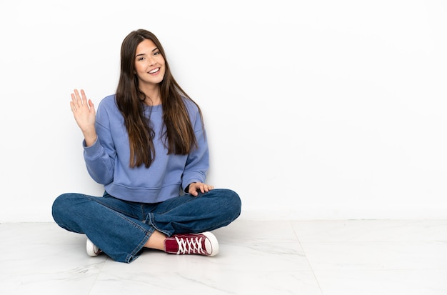 Mujer joven sentada en el suelo saludando con la mano con expresión feliz