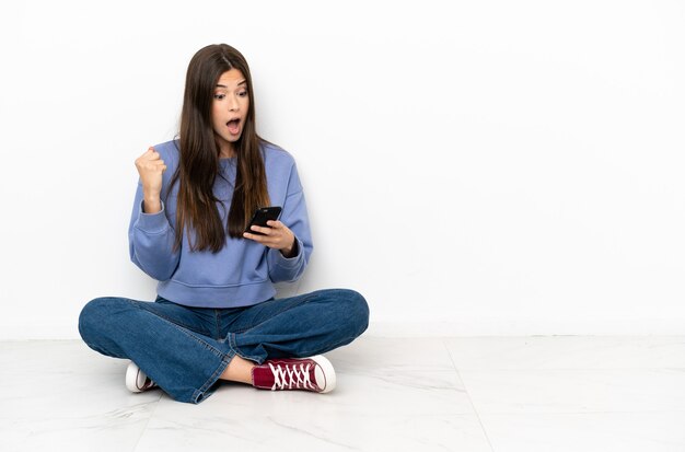 Mujer joven sentada en el suelo sorprendida y enviando un mensaje