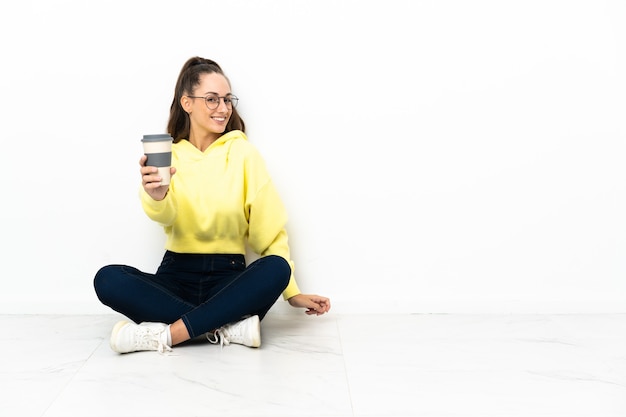 Mujer joven sentada en el suelo sosteniendo un café para llevar con expresión feliz