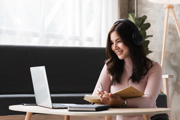 Mujer joven sentada y trabajando en una sala de estar moderna con portátil y portátil con auriculares sonriendo y disfrutando de un ambiente cómodo de oficina en casa