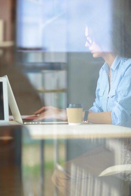 mujer joven, sentado, en, mesa de la oficina, con, laptopview, por, ventana