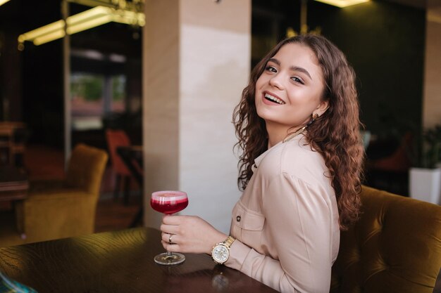 mujer joven, sentado, en, restaurante, y, bebida, cóctel, hermoso, niña, con, pelo rizado, sonrisa, a, cámara