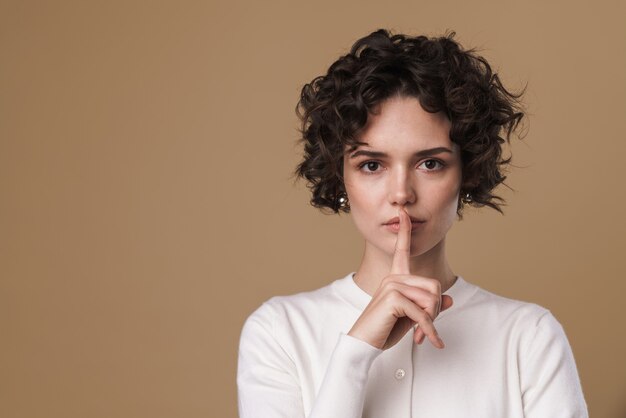 Mujer joven seria posando y gesticulando signo tranquilo aislado sobre pared beige