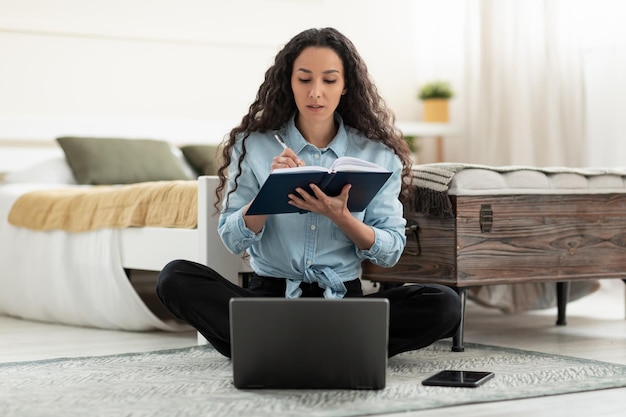 Mujer joven seria que usa una computadora portátil que trabaja en línea tomando notas durante una reunión de negocios desde casa llena