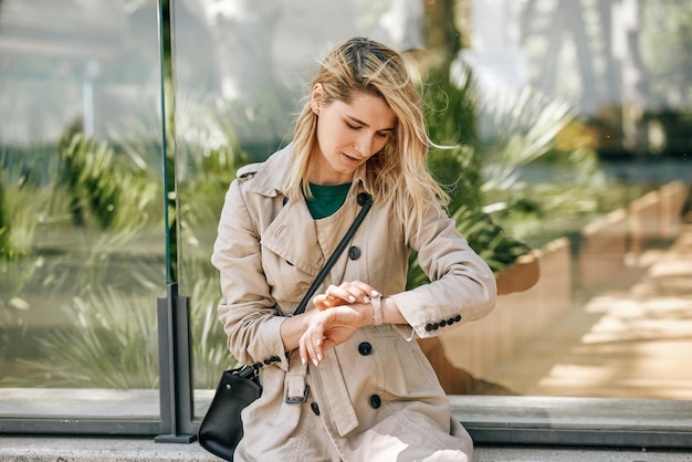 Mujer joven seria sentada al aire libre en la ciudad en un día soleado y esperando a su amiga Hermosa mujer preocupada mirando al reloj esperando a alguien que llega tarde Negocios y estilo de vida de la gente