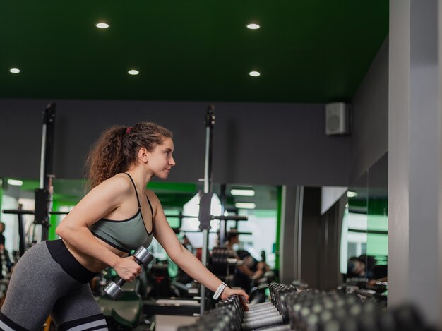 Mujer joven sexy en forma con ropa deportiva haciendo ejercicio con mancuernas de elevación con una mano apoyada en la rejilla en el gimnasio. Entrenamiento con pesas gratis